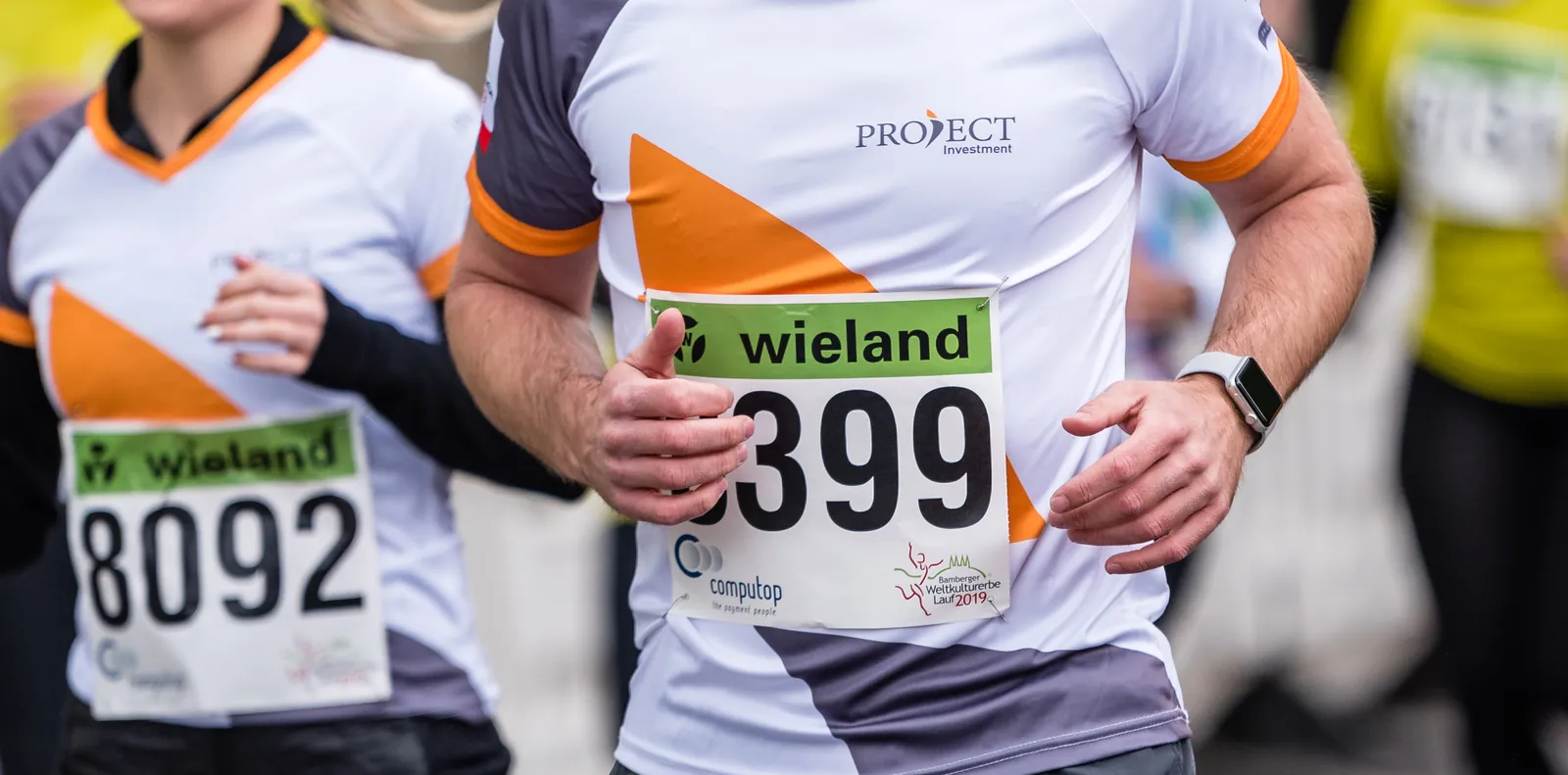 Zwei Läufer in weißen Laufshirts mit orangem und grauem Schwung und PROJECT-Logo, mit Startnummern